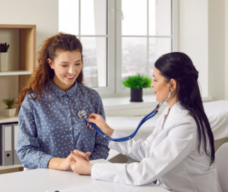 Young woman comes to clinic for heart and lungs checkup. Friendly female doctor sitting at desk in modern medical office, holding stethoscope, listening to patient's breath or heartbeat and smiling