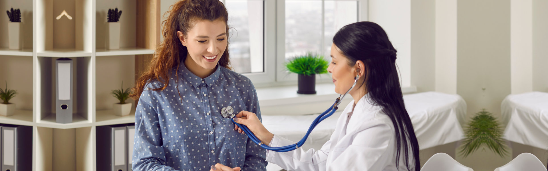 Young woman comes to clinic for heart and lungs checkup. Friendly female doctor sitting at desk in modern medical office, holding stethoscope, listening to patient's breath or heartbeat and smiling